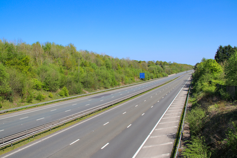 A photograph a three-lane motorway in Britain, bordered by tree-lined embankments and without any vehicles in sight.